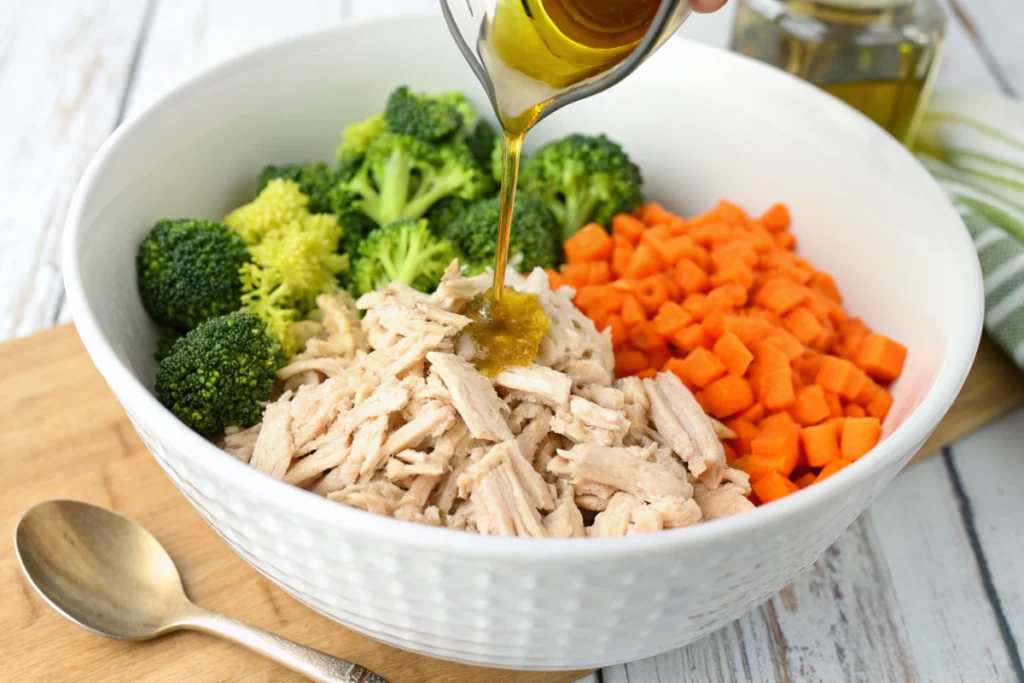 Shredded rotisserie chicken with vegetables and olive oil in a mixing bowl.