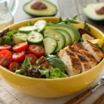 A yellow bowl filled with a healthy salad consisting of grilled chicken, avocado, cucumber, cherry tomatoes, and mixed greens.