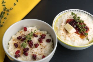 Two bowls of creamy rice pudding topped with dried cranberries, thyme sprigs, and spices are presented on a dark surface.