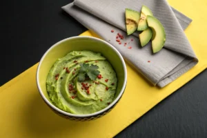 A bowl of vibrant green guacamole topped with red pepper flakes and a sprig of cilantro sits on a yellow placemat.