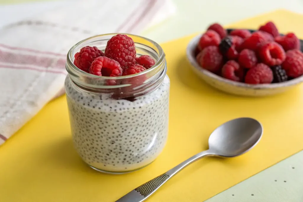 A jar of chia seed pudding topped with fresh raspberries sits on a yellow surface next to a spoon and a bowl of berries.