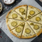A white pizza topped with pickle slices and herbs sits on parchment paper.