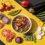 A bowl of hearty soup sits on a yellow and black background surrounded by fresh vegetables.